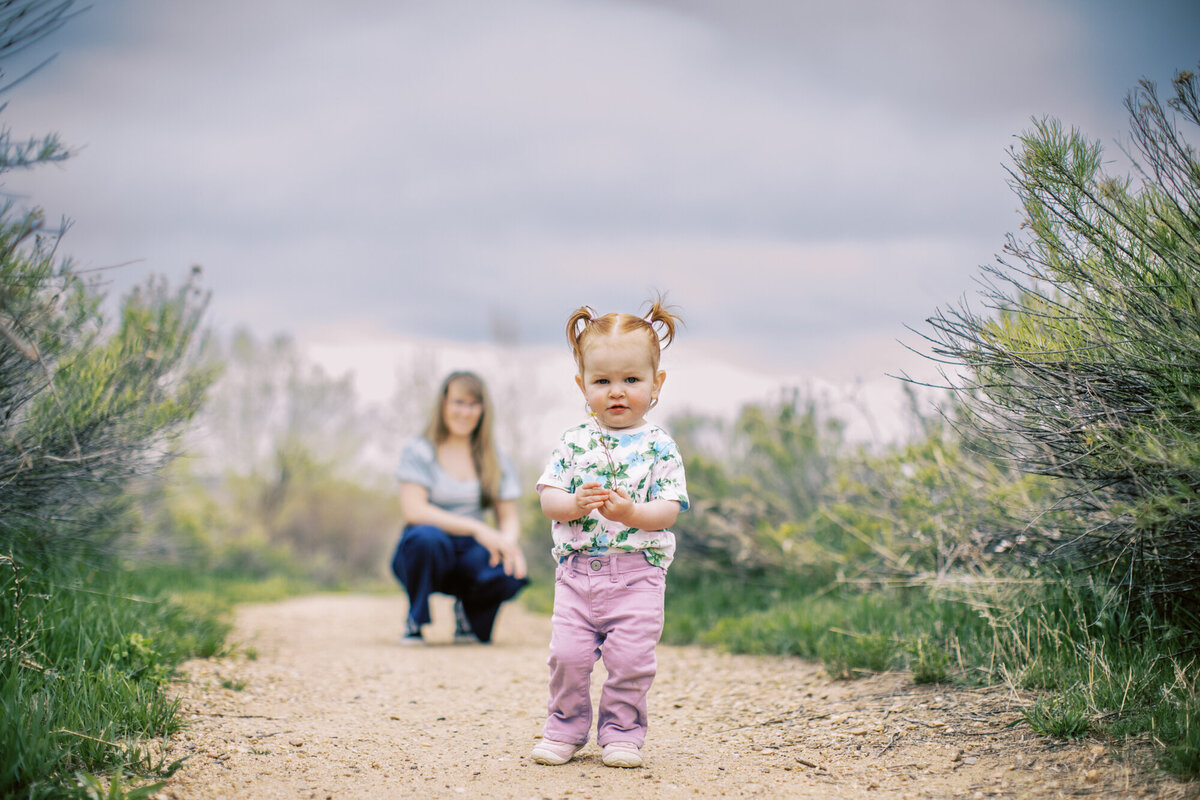 toddler girl with pink pants and pigtails stands in front of her kneeling mom
