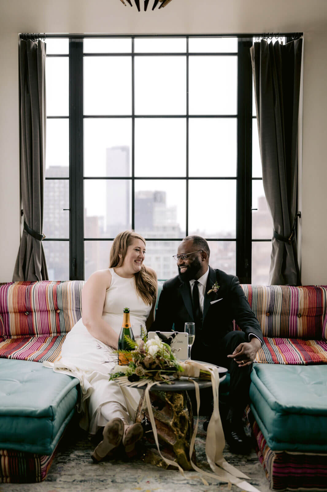 The bride and the groom are happily seated on a couch with a bottle of champagne inside a Ludlow Hotel room in NYC.
