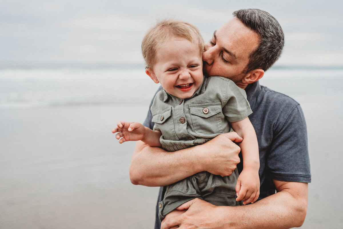 Father holding and kissing young daughter who is laughing at Del Mar beach, San Diego family photographer.