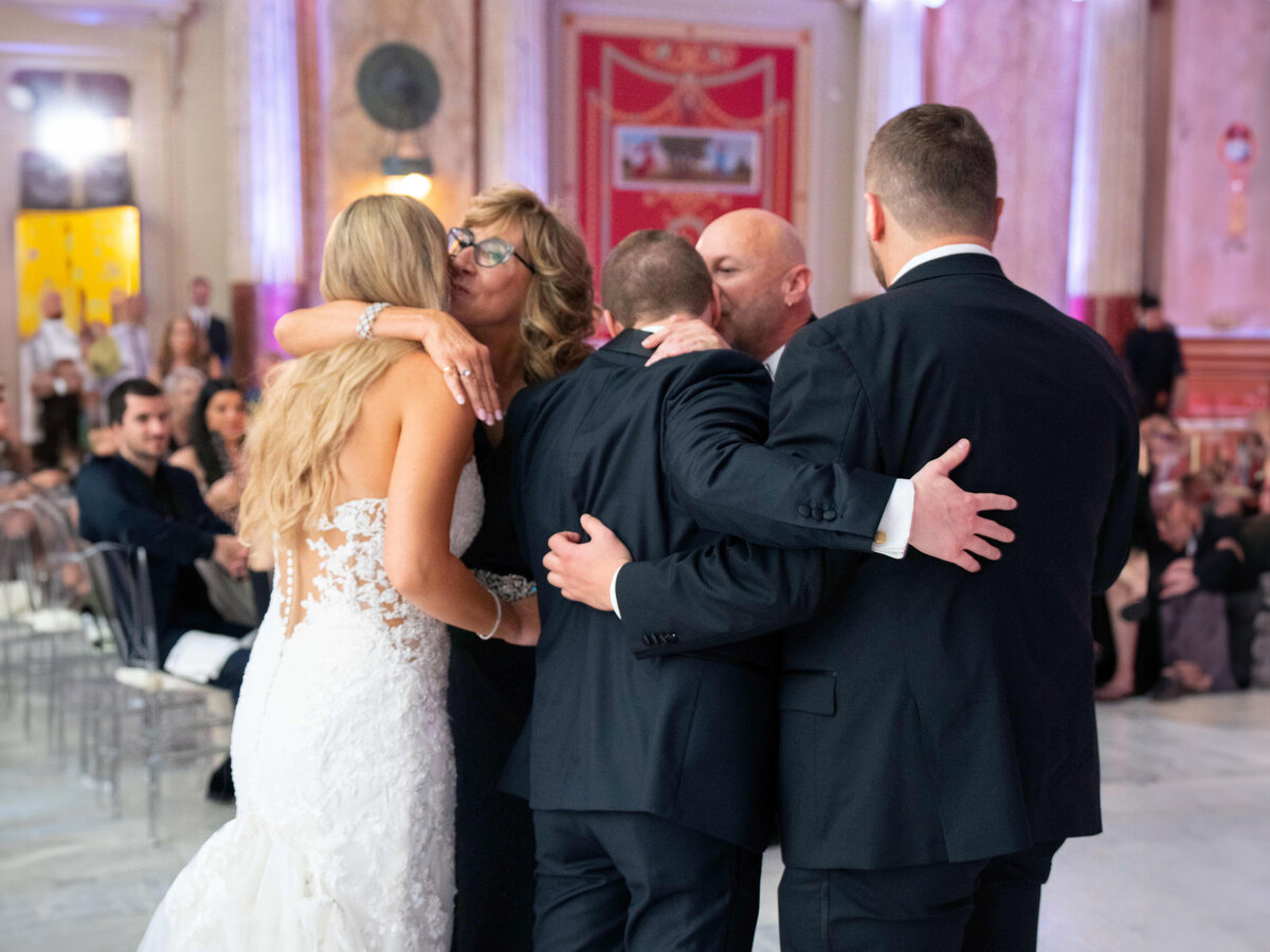 A bride in a white dress and three men in suits embrace a woman wearing glasses during a wedding celebration in an elegant venue, with onlookers sitting in the background.