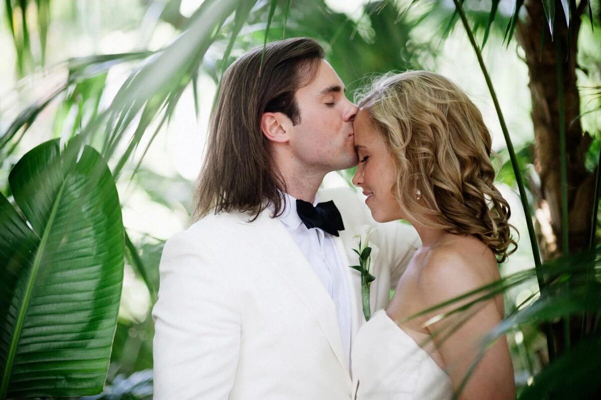 A bride and groom share a tender kiss amidst lush greenery, enveloped in the natural beauty of their surroundings.