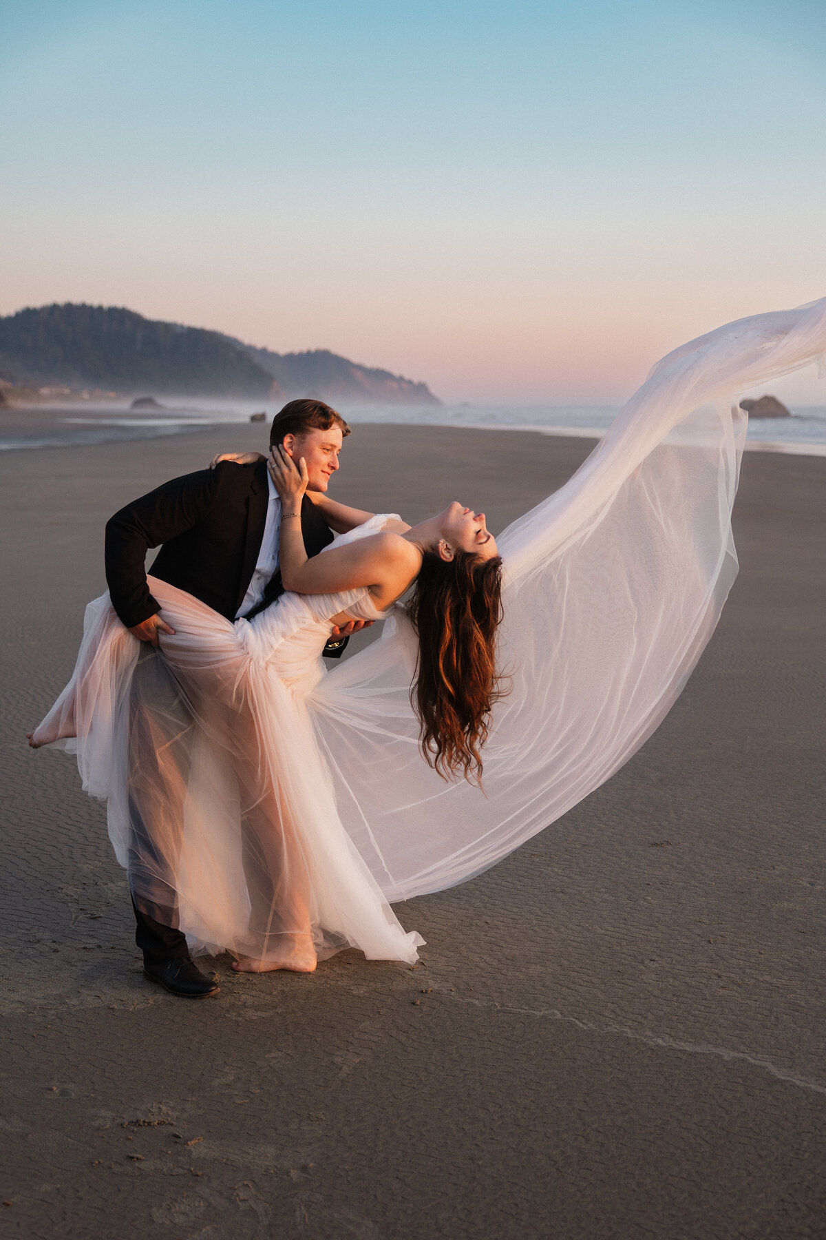 Groom dips bride with her dress flowing in the wind on the beach.