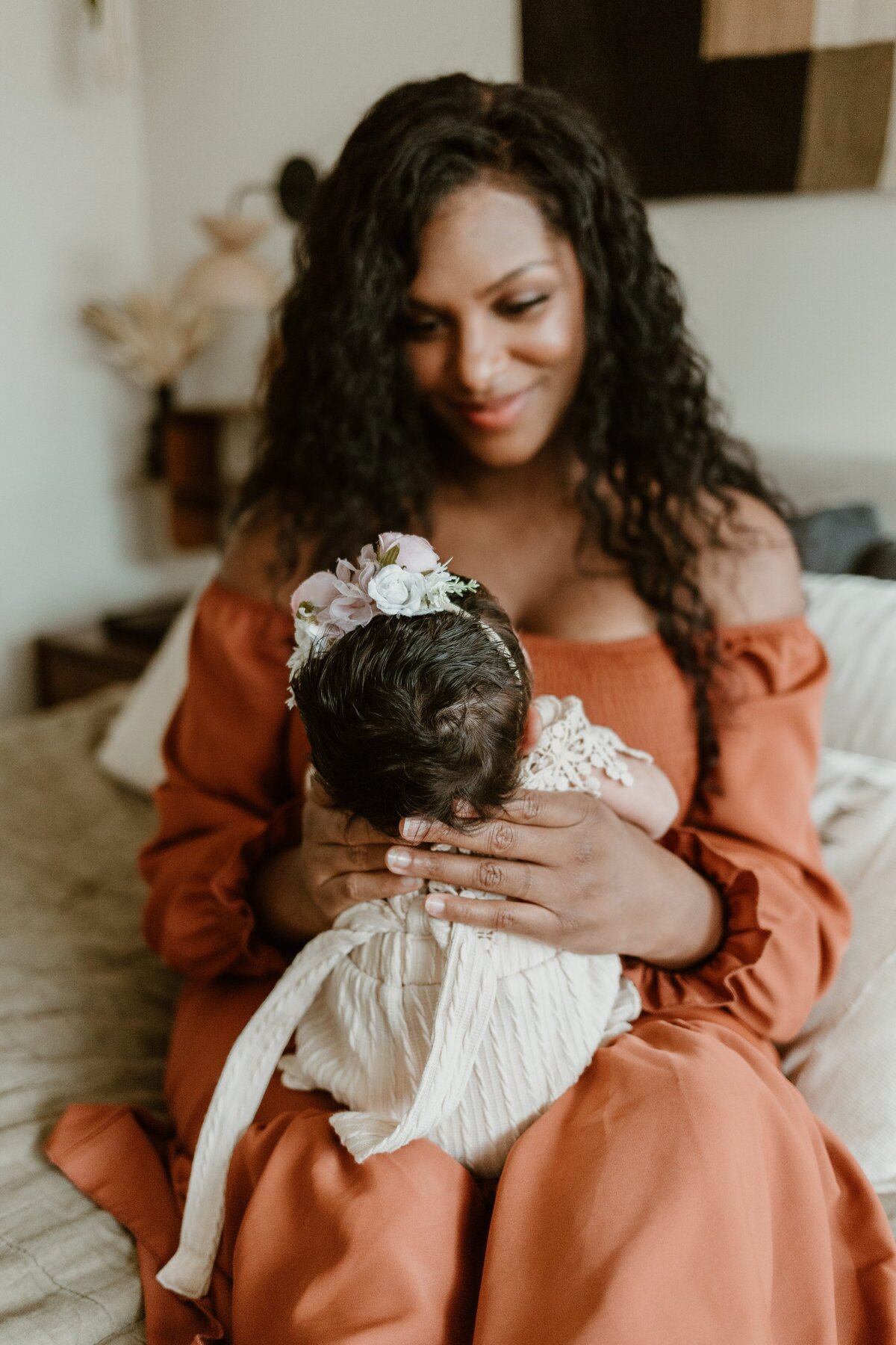 A Hudson Valley newborn photographer captures a tender moment as a mother in a burnt orange dress smiles down at her baby adorned with a flower crown, nestled comfortably in her arms.