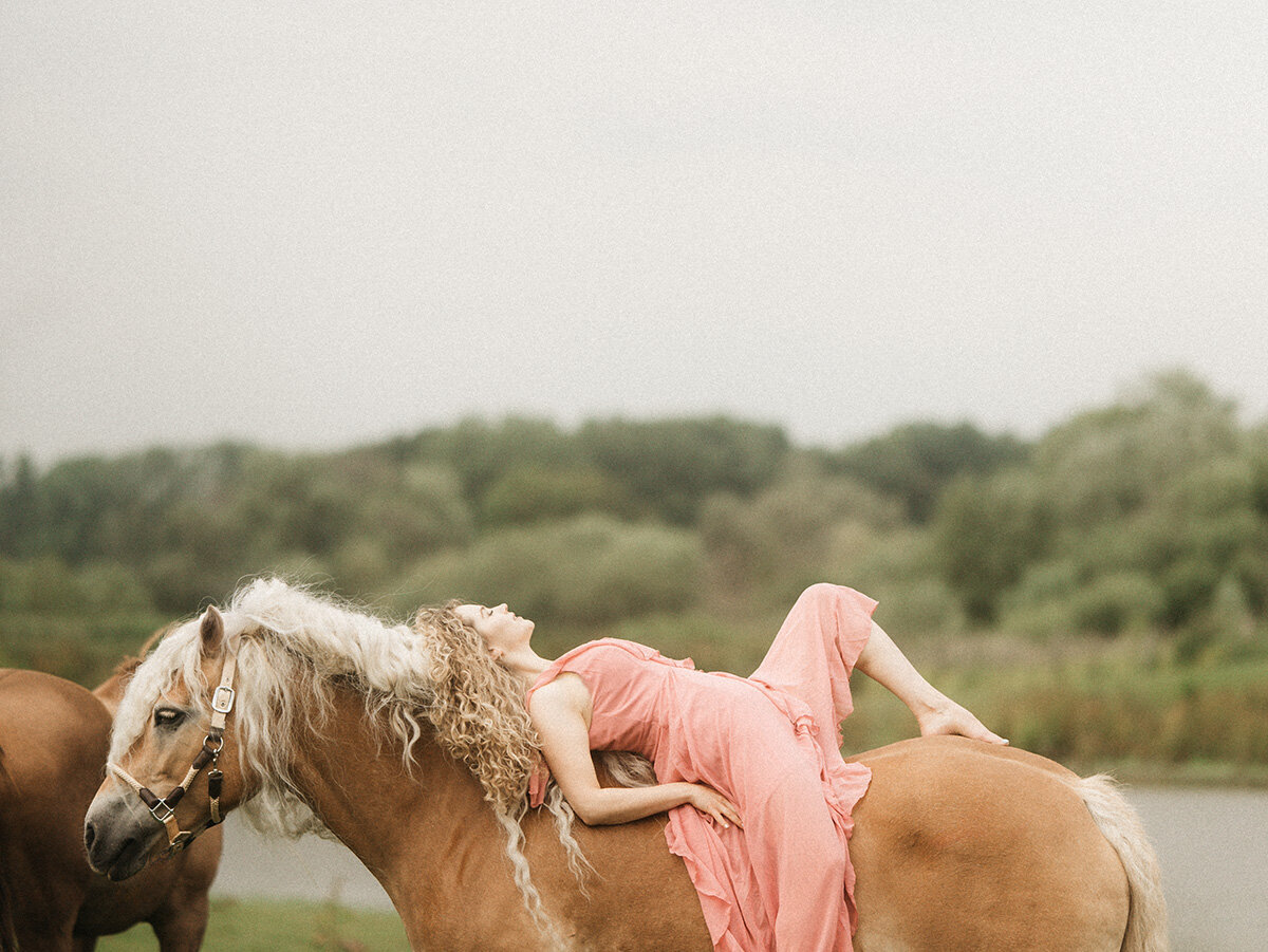 Naomi ligt in een roze jurk op haar Haflinger Storm, met haar ogen gesloten, omgeven door groen – een zacht, filmisch portret van de pure band tussen mens en paard.

