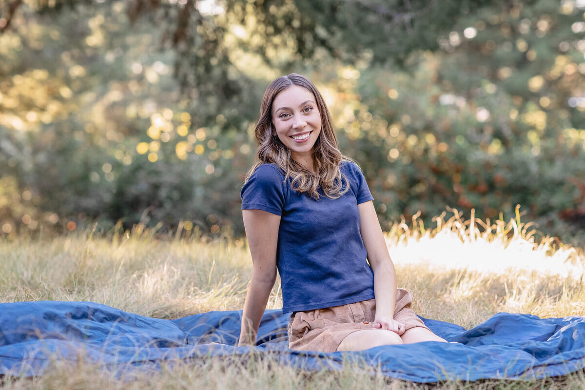 A young woman with long, brown hair in a navy shirt sitting on a blue blanket in green grass at Jeffrey Open Space.