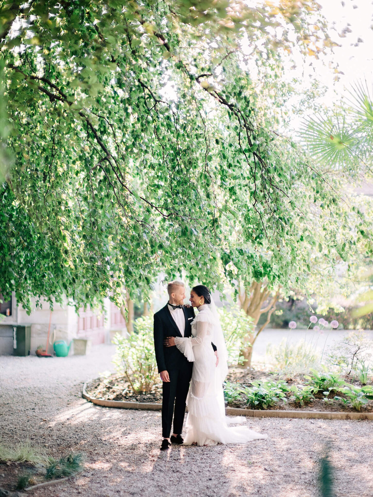The bride and groom are standing in a garden with plants and trees in the background.