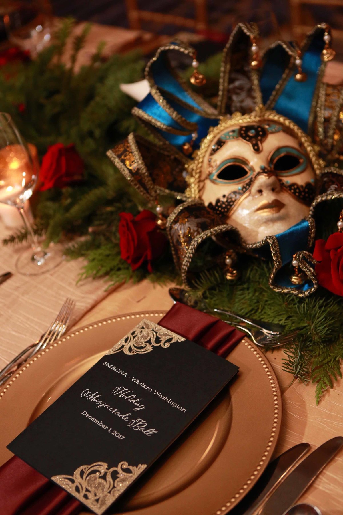 holiday party place setting with red napkin, black menu card on gold charger plate and evergreen garland runner and vintage carnival mask