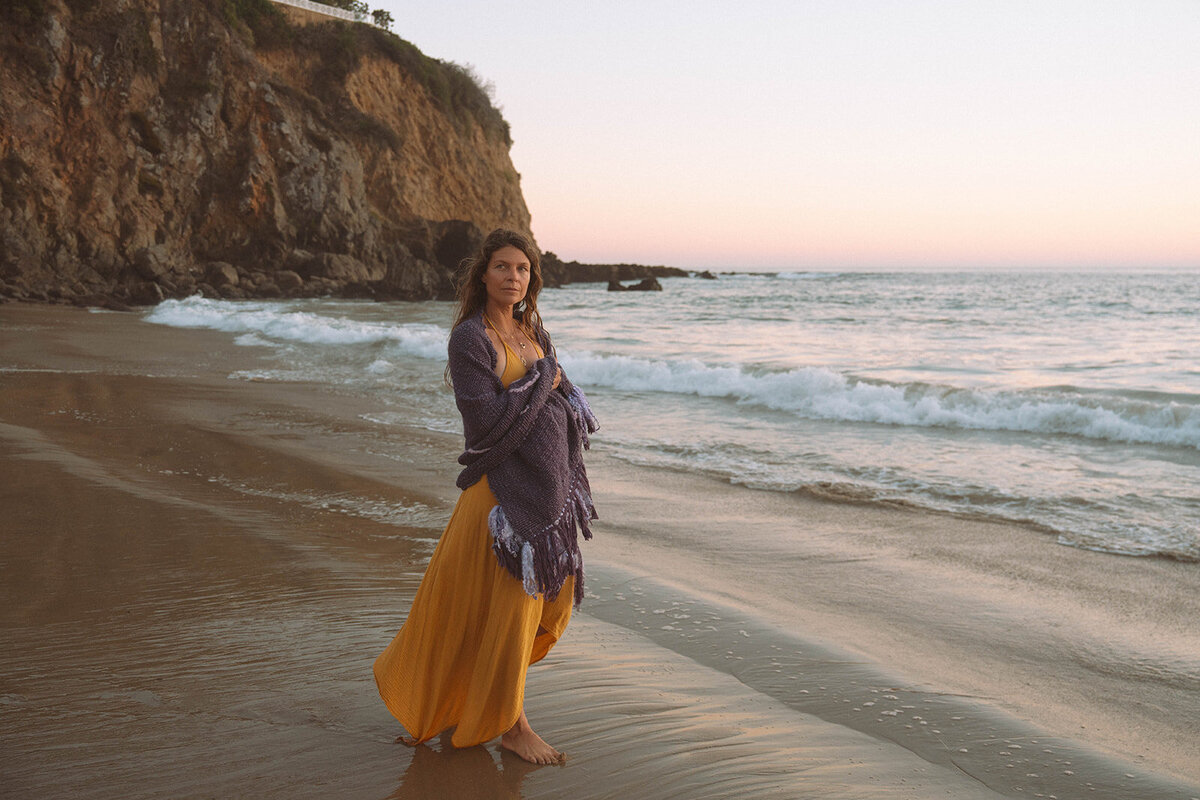 Branding photo of a woman walking along the shoreline in Laguna Beach at sunset, wearing a yellow dress and purple shawl. Editorial portrait by Orange County photographer Maria Alcantara.