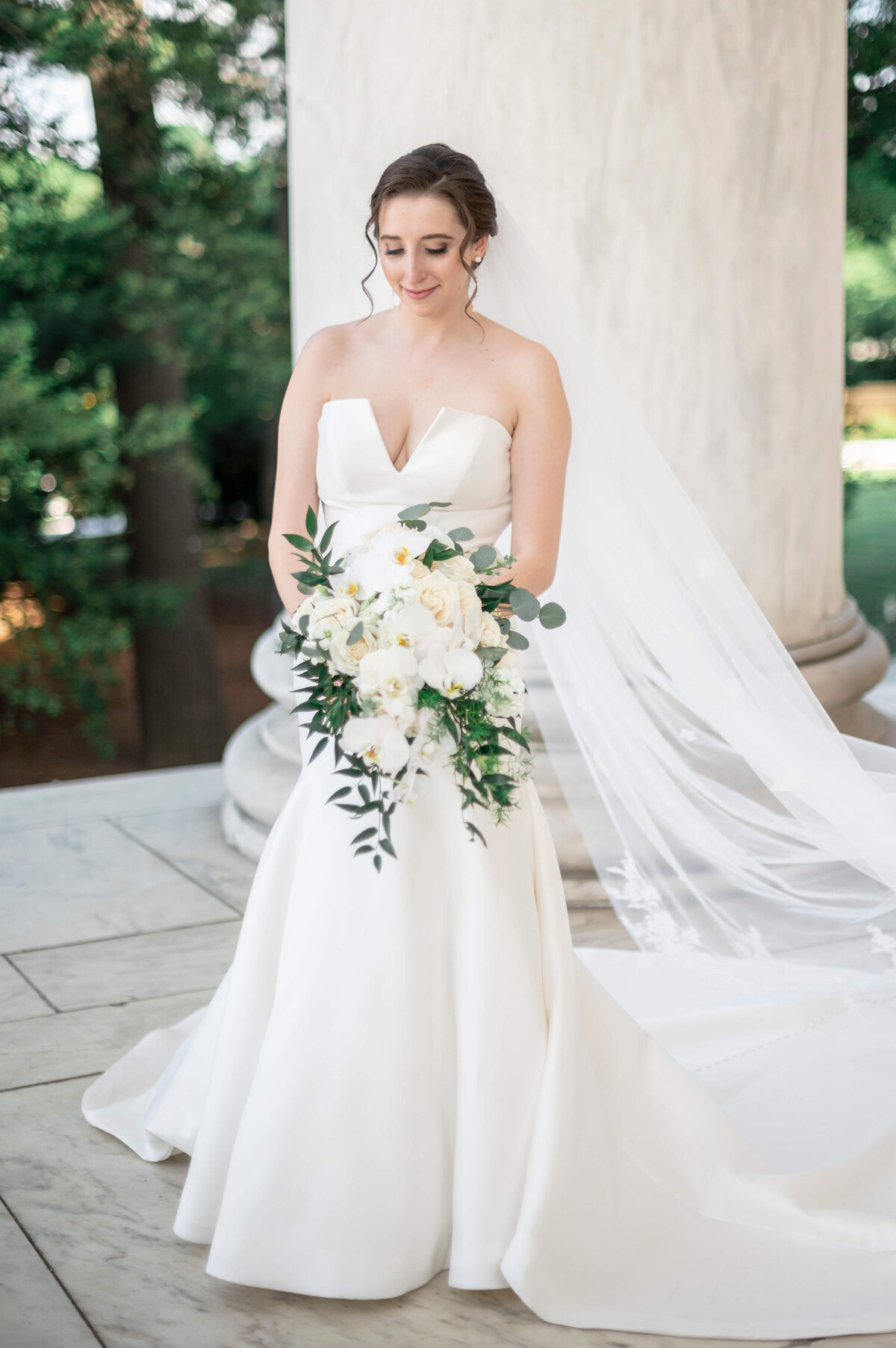 A bride stands in a strapless white gown with a long veil, holding a bouquet of white flowers and greenery. She is outdoors in front of a large column, with trees visible in the background. Her gaze is directed downwards.