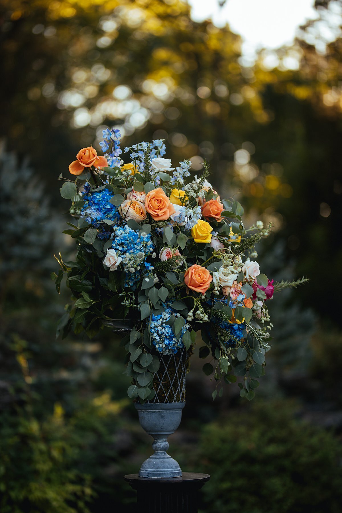 A large gray urn of flowers for a wedding ceremony at Cheekwood Botanical Gardens. The stone urn has blue, orange and ivory roses with an assortment of greenery and accents of hot pink flowers.