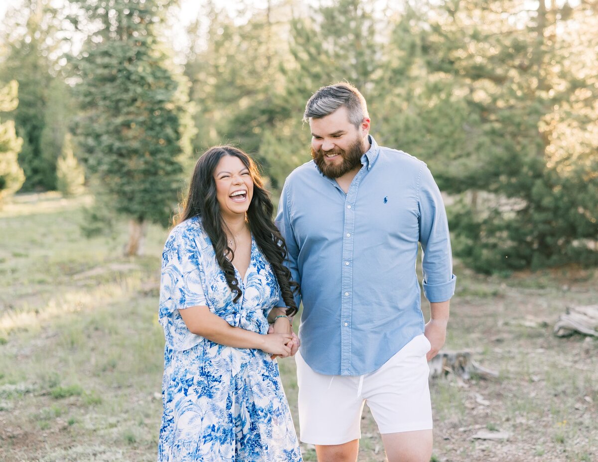 A young couple, dressed in a blue dress and blue shirt are walking hand in hand among the pine trees. They are seen laughing together and having a great time during their engagement session. 