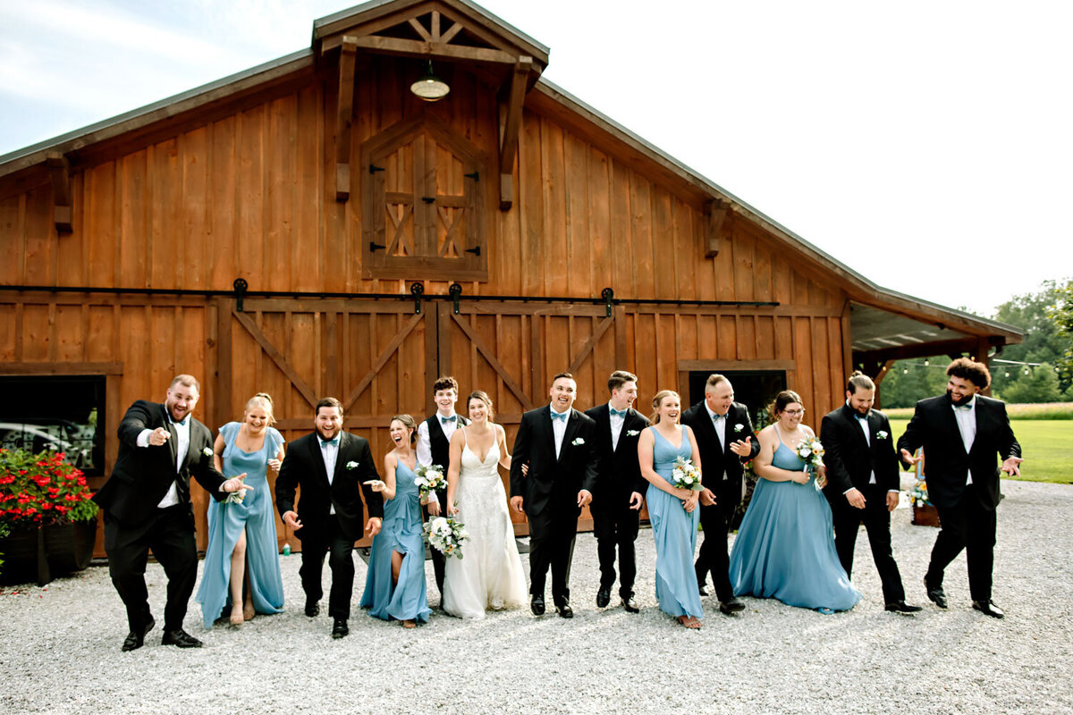 Wedding party in blue and black attire standing in front of a wooden barn, smiling