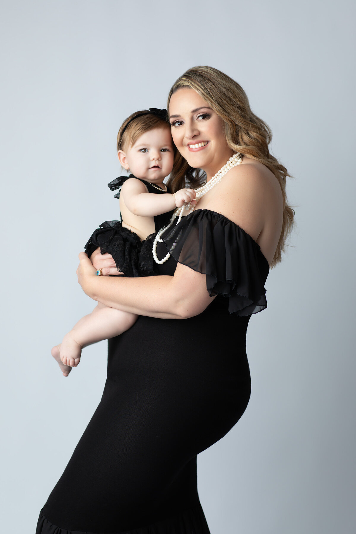 Dallas milestone photographer showcases a glamorous mother and daughter dressed in coordinating black outfits and pearls, capturing a joyful moment full of connection and elegance.