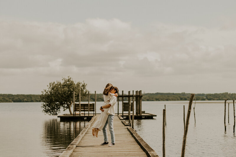 Homme soulevant sa chéri dans ses bras sur un ponton en bois. Décor de lac et de nature en arrière plan. Capturés par Laura, photographe couple en vendée.