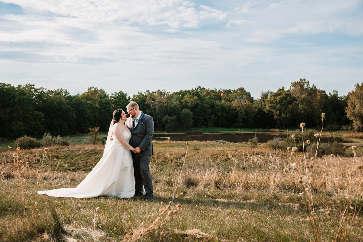 A bride and groom smile at each other while snuggling in a field by a pond