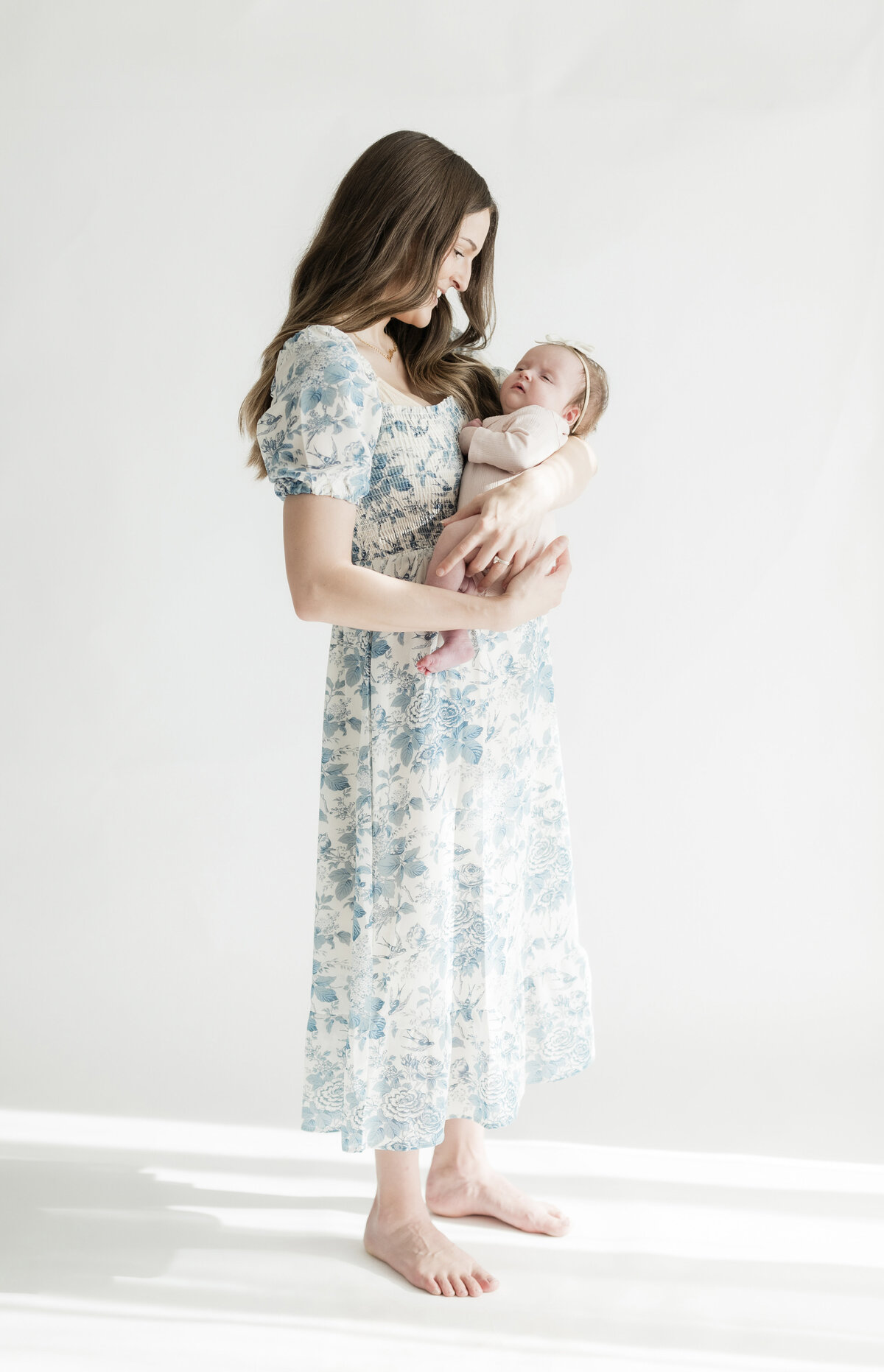 Mother in a floral dress standing barefoot, holding her baby in front of a clean white backdrop.