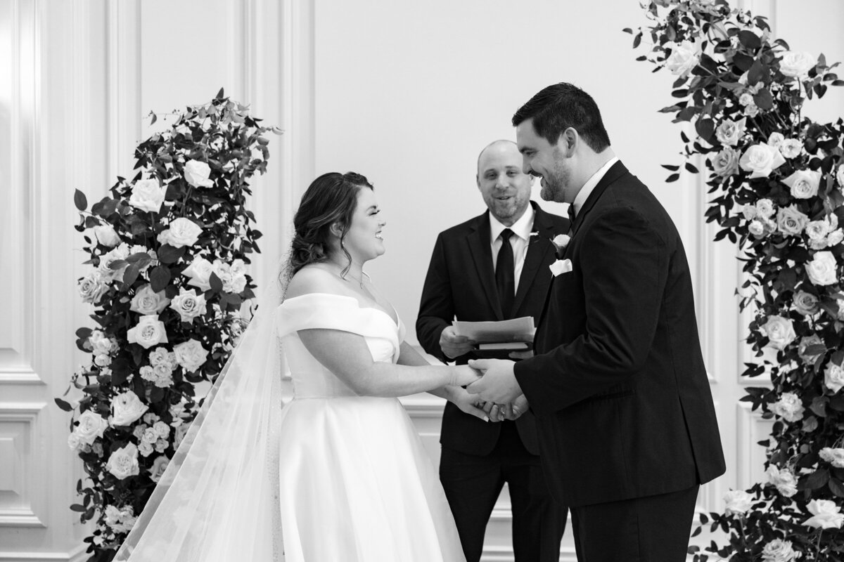black and white photo of the bride and groom holding hands and laughing during their ceremony in the Governor’s Room at The Adolphus in Dallas, capturing a joyful and timeless wedding moment.