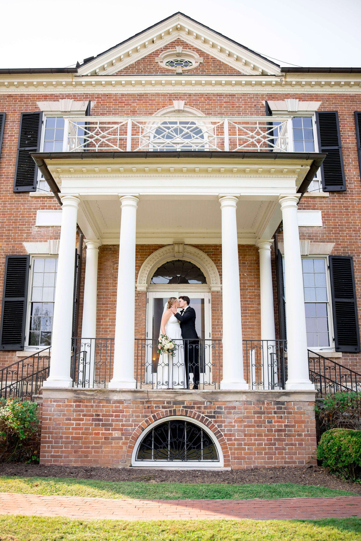 A couple in wedding attire shares a kiss under a grand, columned entrance of a two-story brick building with black shutters and white trim. A semi-circular window is above them. Greenery surrounds the entrance.