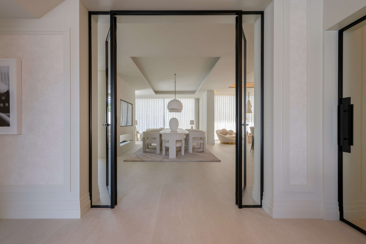 Through open glass doors, an entrance to a dining area with a large table and pendant lights. The room has a bright, airy aesthetic with natural light coming in from large windows behind the table.