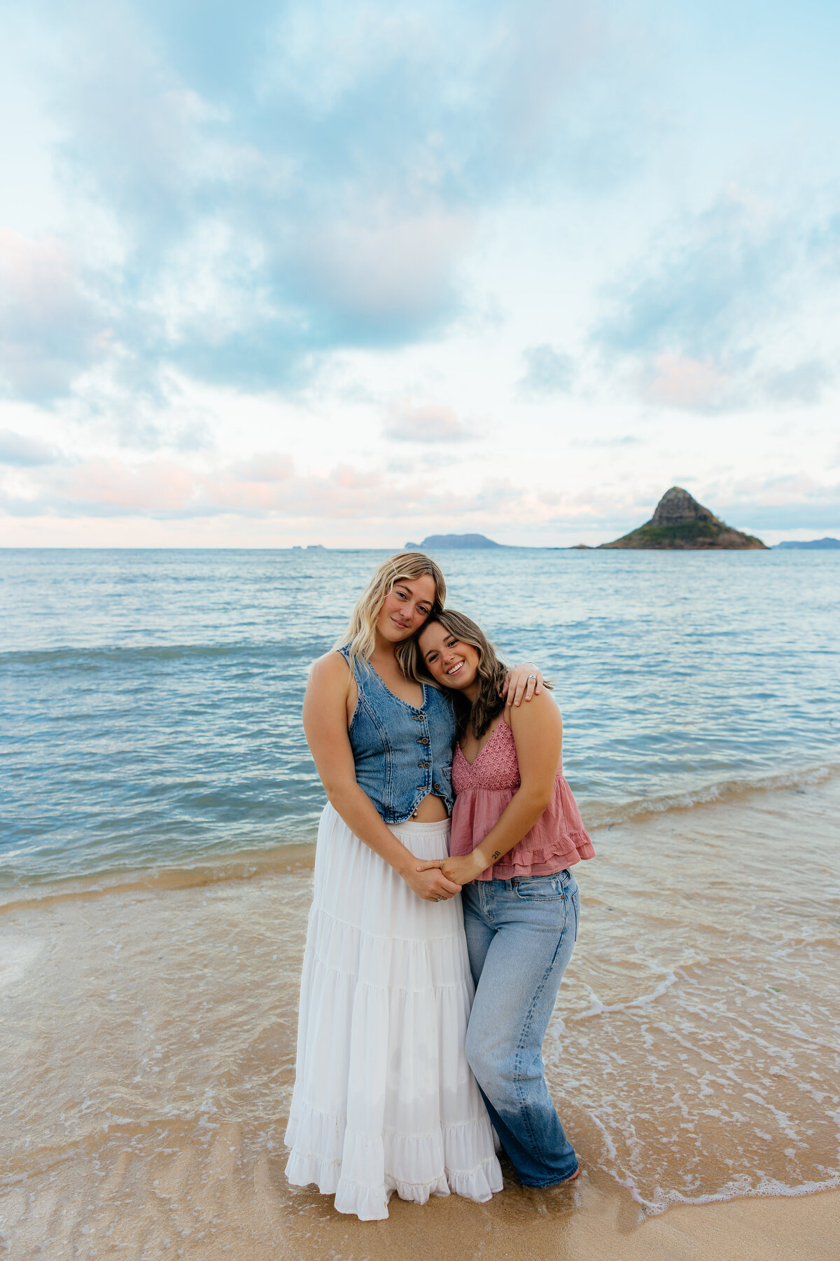 Two best friends smiling and hugging at the edge of the ocean during a sunset photoshoot in Oahu, Hawaii, with a small island in the background.
