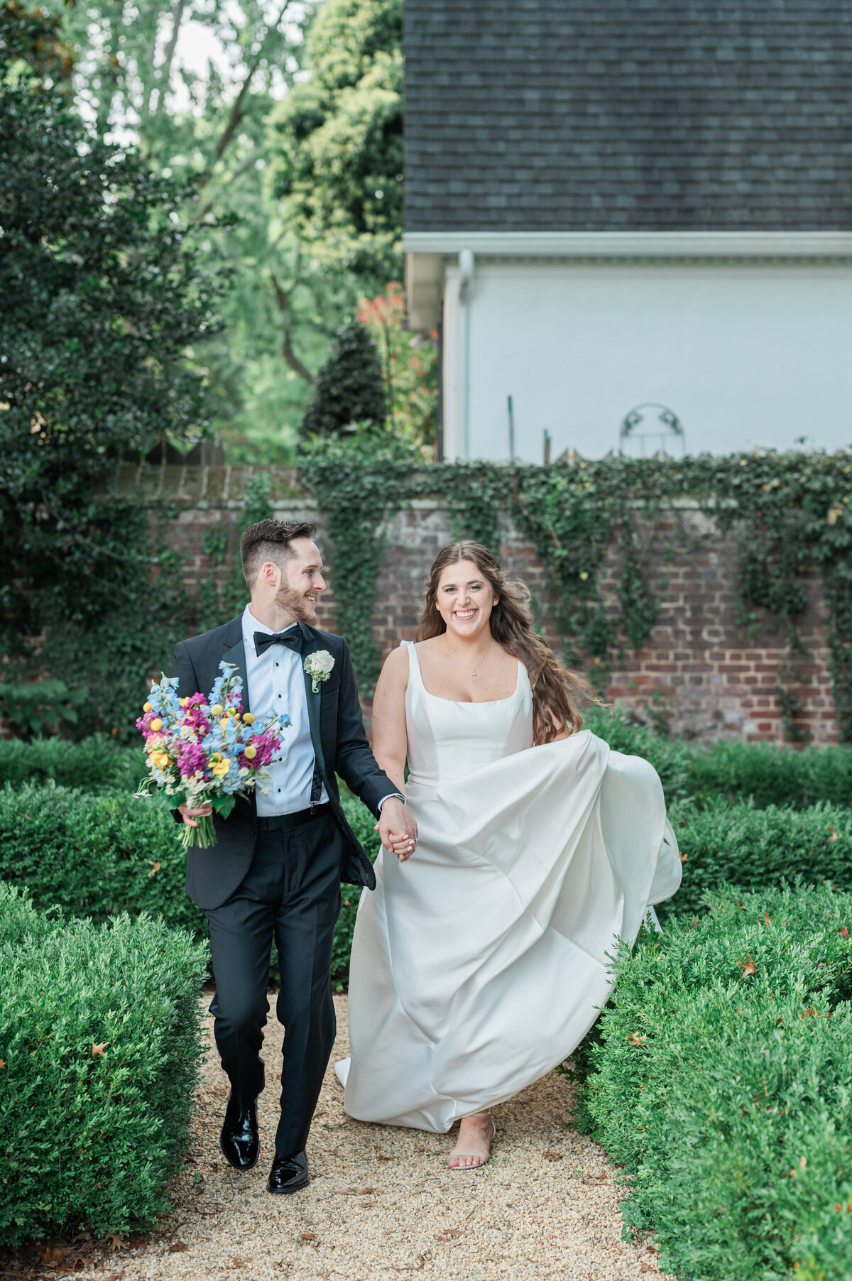 A wedding couple walking through a small garden while one holds the bottom of their dress and the other holds a bouquet