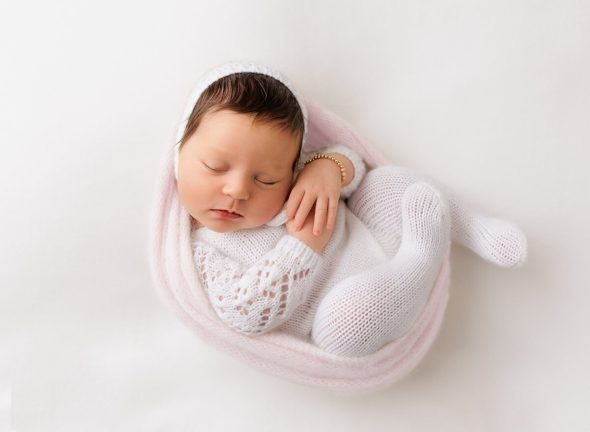 Baby girl in a white knit onesie is curled in a pink swaddle with her hands folded atop of her chest. She is sleeping peacefully. Captured by premier Brooklyn NY family photographer Chaya Bornstein Photography.