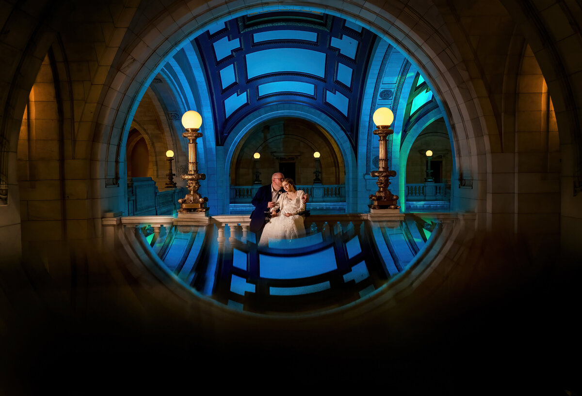 A bride in a wheelchair and her husband hold each other. The Old Courthouse lights and ceiling are mirrored into a circle.