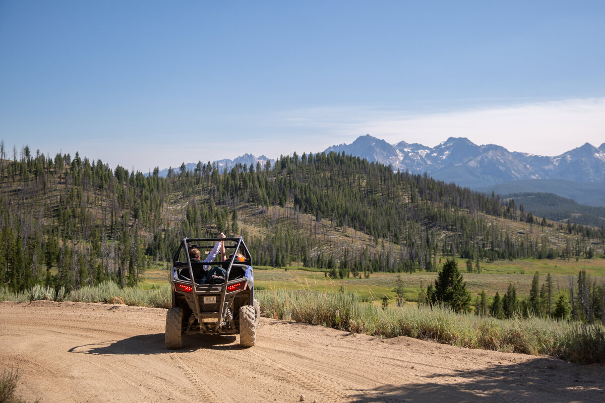 A side-by-side is parked on a dirt road in Idaho and the couple inside of it are holding hands and raising them in the air.