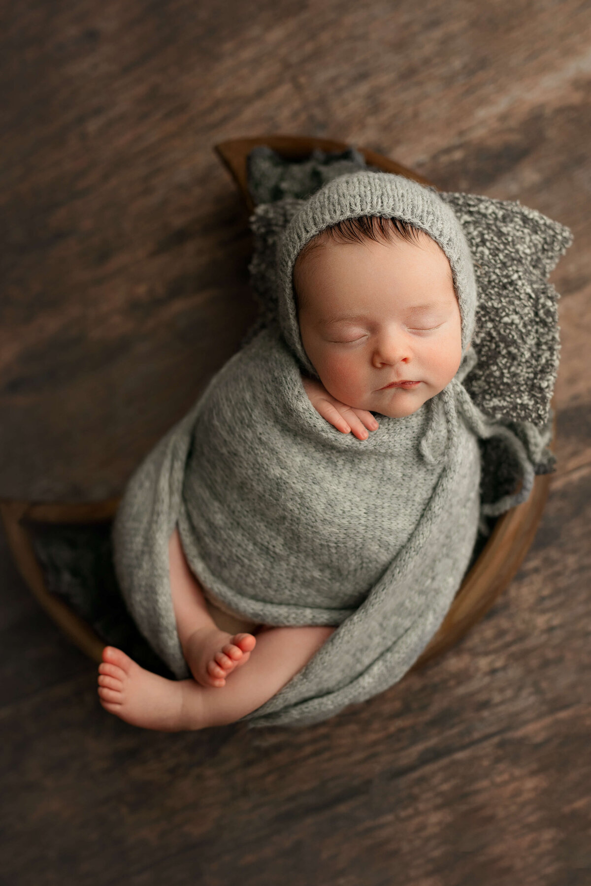 newborn baby boy in a grey wrap and bonnet laying on a pillow in  a wooden moon bowl