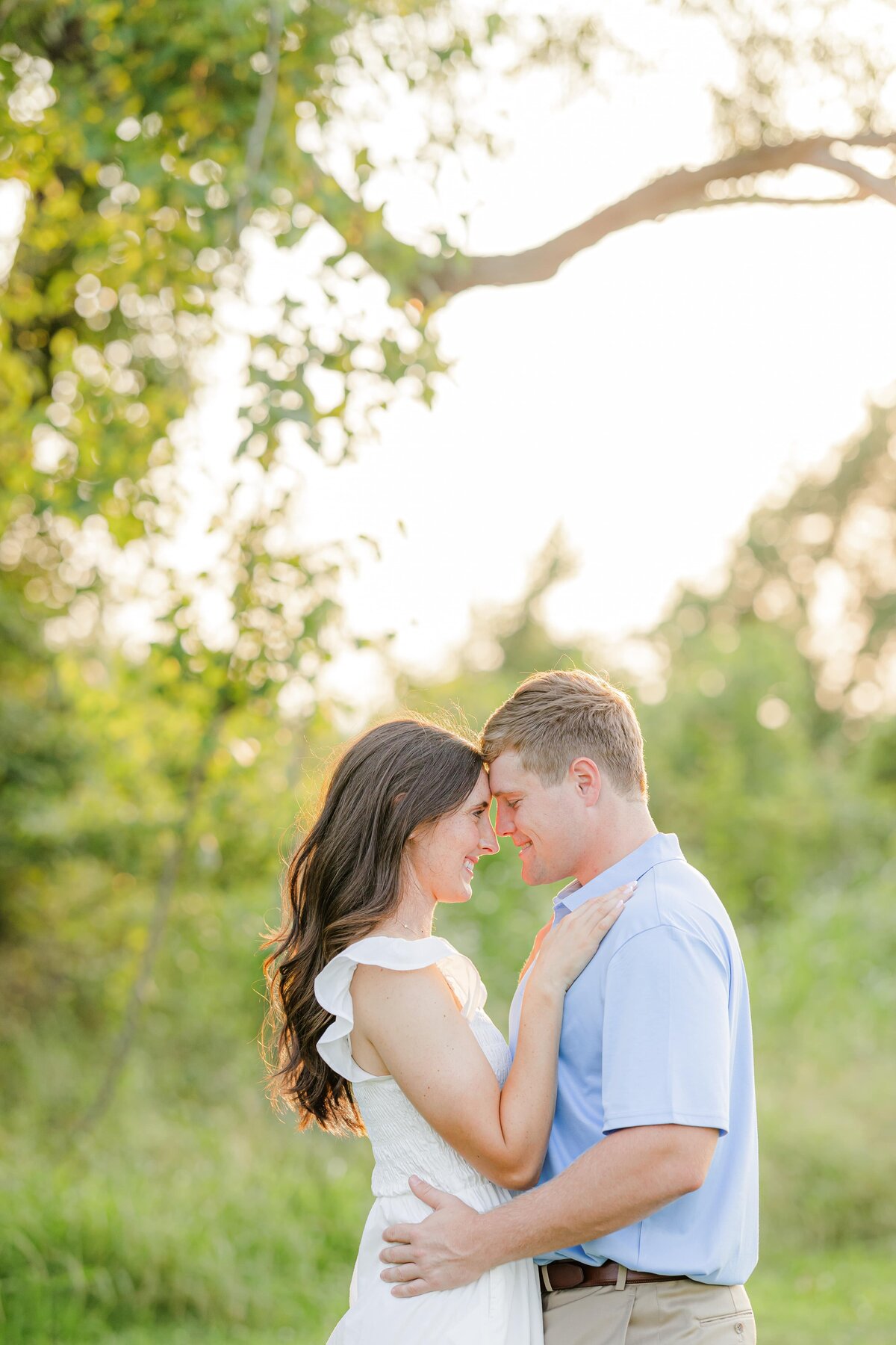 A woman in a white dress hugs and touches foreheads with her fiancee in a field at sunset