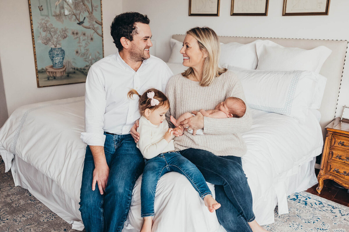 mom and dad hold their beautiful daughters while they sit on their bed smiling at each other in their Del Mar home in san diego