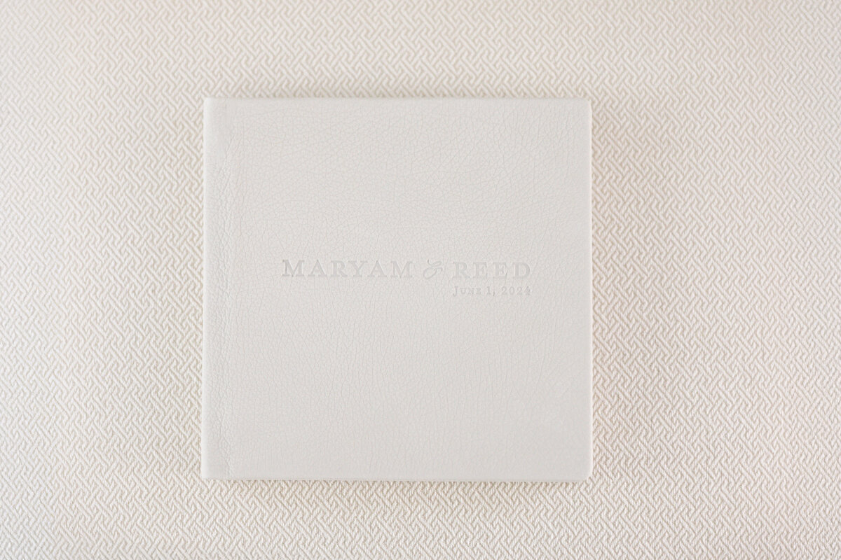A white photo album cover embossed with Maryam & Reed, June 3, 2014 is placed on a textured surface.