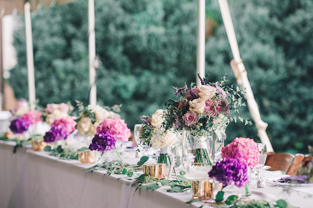 Head table at a tented wedding at Rippavilla Mansion with a white tablecloth decorated with gold votive candles, pink and purple hydrangea and the bridal bouquet.