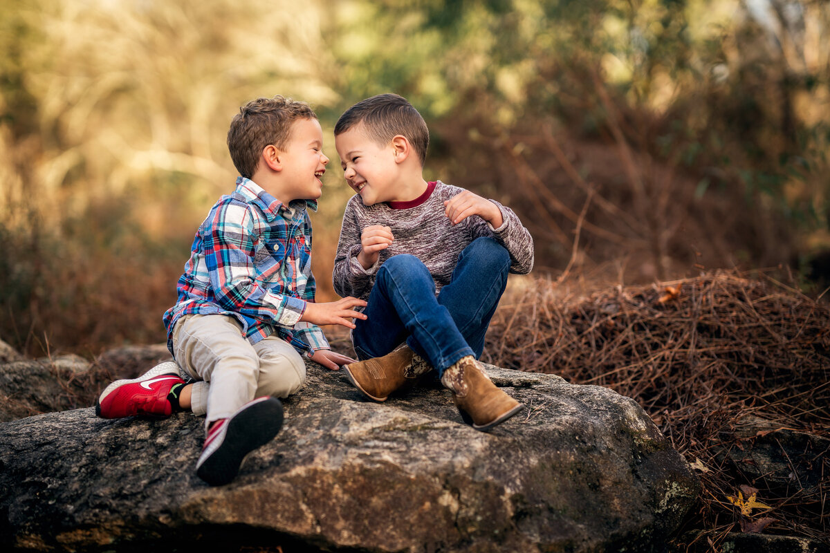 Two young brothers are sitting on a rock and laughing at each other.