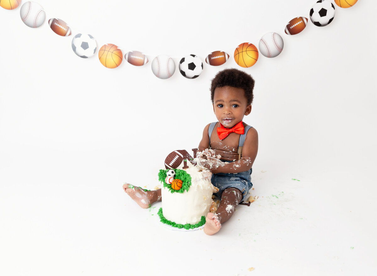 Baby boy sits with a sports themed cake between his legs for his first birthday cake smash photoshoot. Baby has icing covering his arms and legs. Baby is looking at the camera.