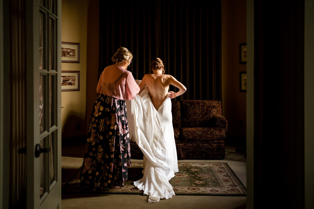 A bride in her wedding dress is silhouetted against a window, being helped by a bridesmaid in a room with classic decor.