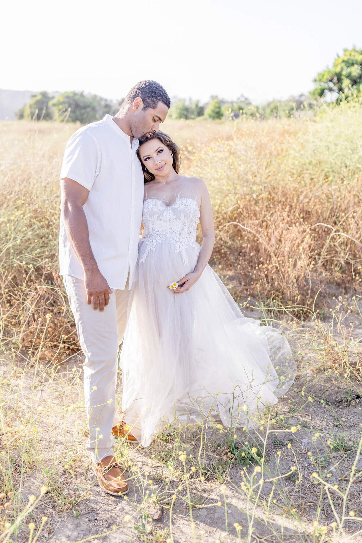 A couple stands in a sunlit field. The woman, wearing a flowing white dress, rests her head on the mans shoulder while gently holding a flower. The man, dressed in a light shirt and pants, stands close, embracing the intimate moment.