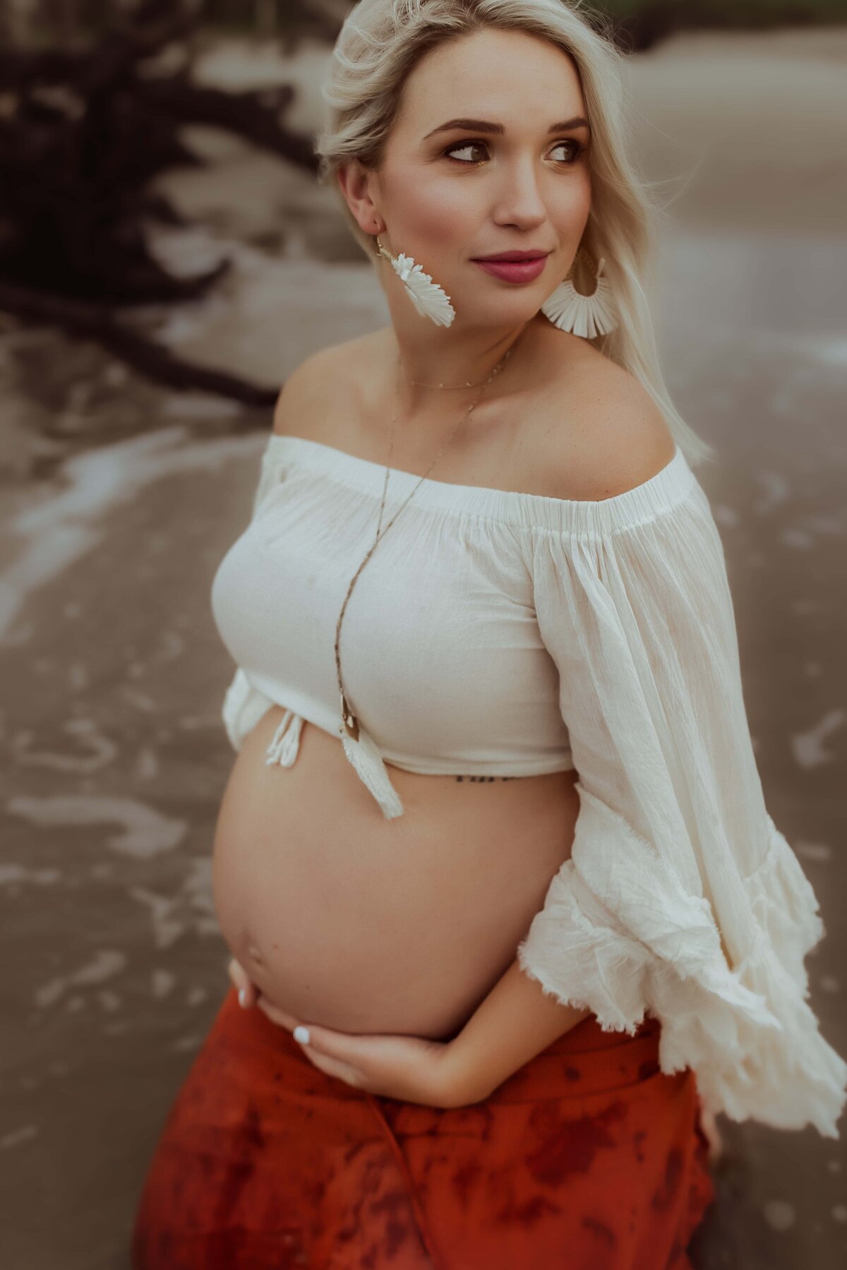 A pregnant woman in a white off-shoulder top and red skirt sits in shallow water on the beach, with one hand supporting her belly.