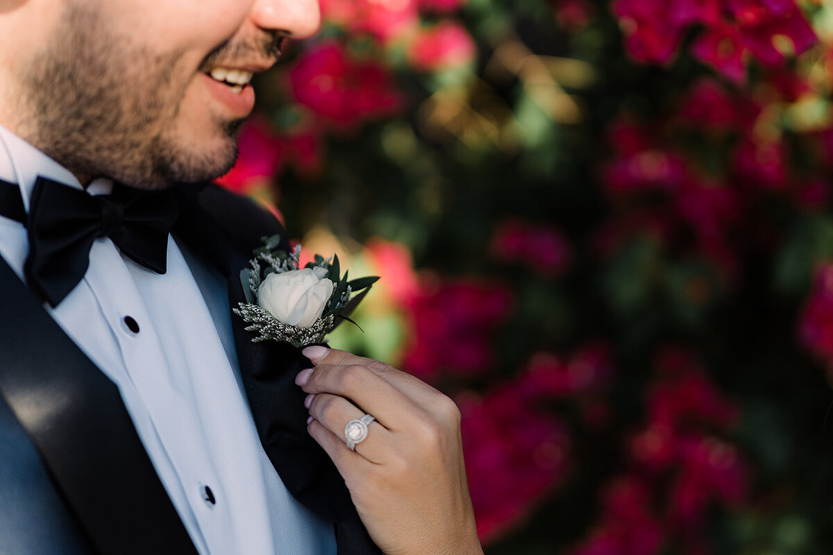 Close-up of the groom's boutonniere, a small white rose with greenery, as the bride’s hand gently adjusts it, with her engagement ring visible.