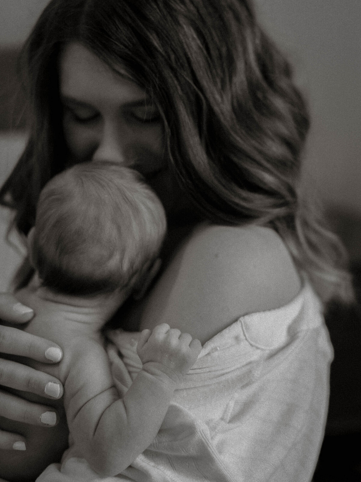 Black and white photo of a mother holding her diapered newborn facing her, gently smelling or kissing his face during a quiet in-home session filled with connection.