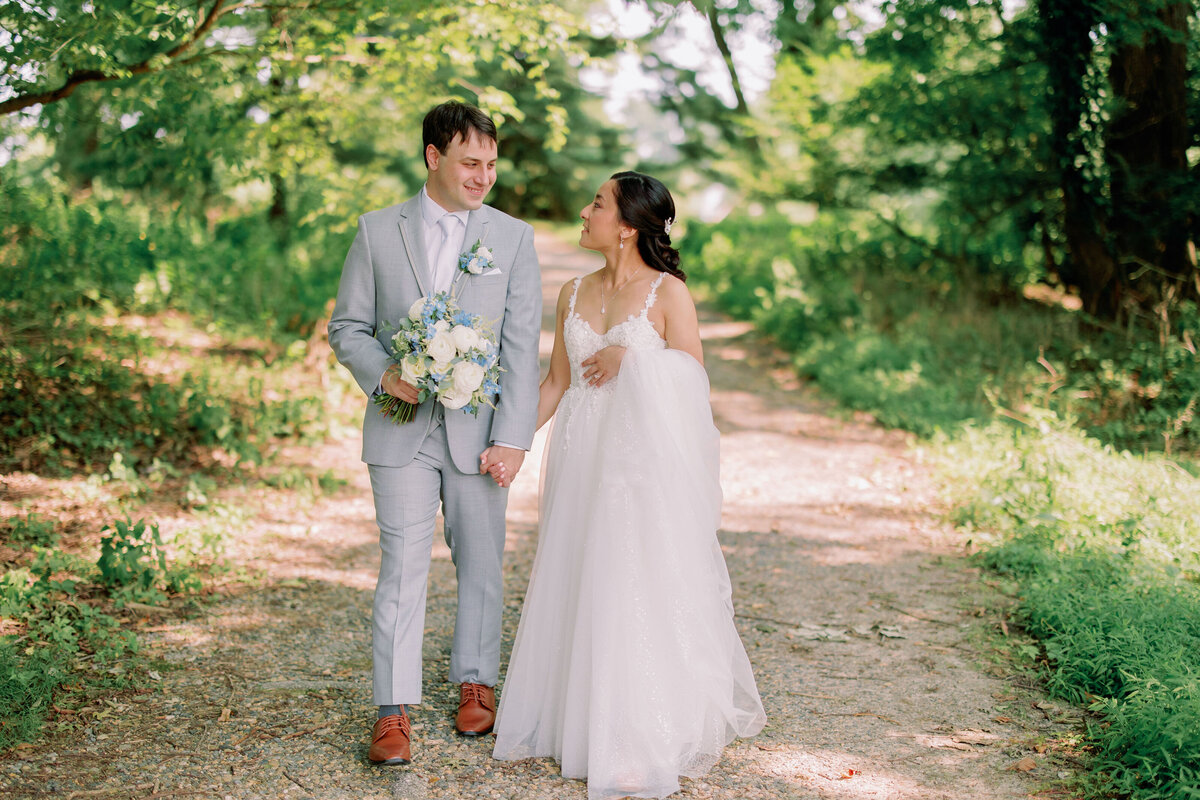 A newlywed couple holding hands and walking along a path as they smile 