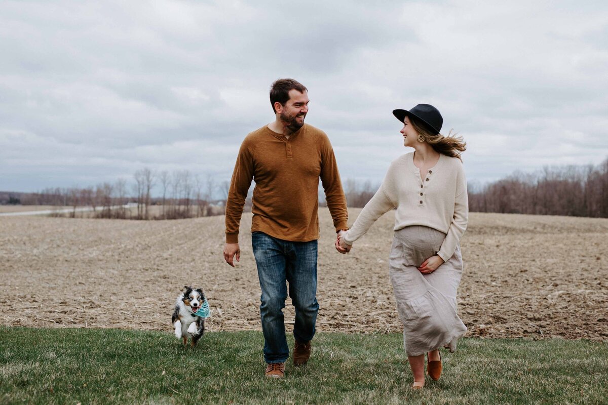 Maternity photoshoot in Exeter, Ontario rustic farm. Mom and dad are walking hand-in-hand smiling and looking at each other. Mom's other hand is under her bump. Their small dog is walking beside them.  The farmers field has been plowed as it is late fall.