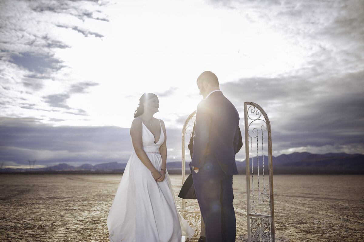 Dry lake Bed elopement Blue Suit on Groom  flowers by michelle  bride in cream color wedding dress with deep  plunging  neckline mountain skyline  sunset las vegas wedding photographers mk delacy photography sunflar photography elvis impersonator