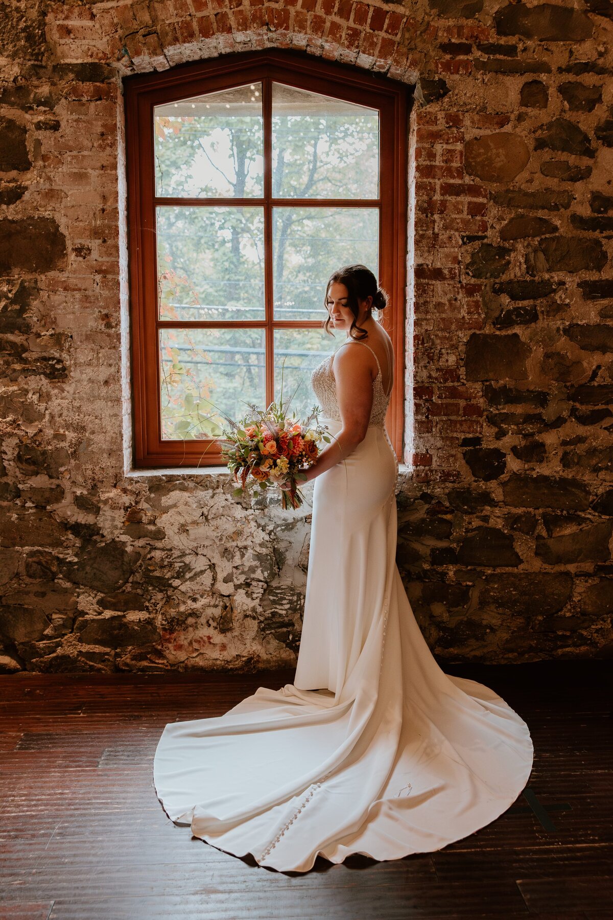A bride poses gracefully in front of a large rustic window at West Hills Country Club. She wears a fitted gown with a long, elegant train, holding a vibrant bouquet of flowers. The warm natural light from the window highlights her serene expression, while the textured stone wall adds a timeless, romantic backdrop to the scene.
