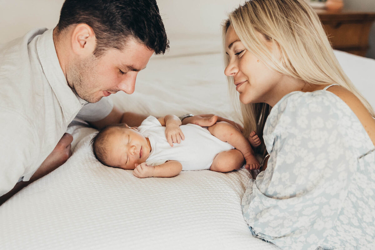 father and mother look at newborn daughter while she lays on a bed in a white onesie