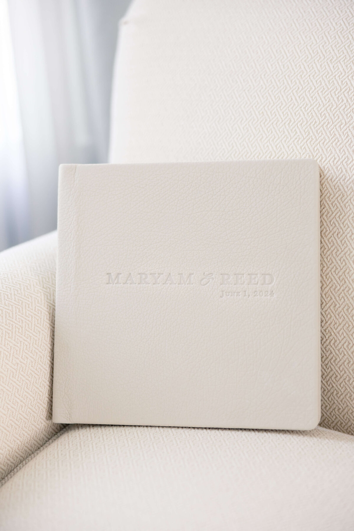 A beige leather photo album with embossed text sits on a patterned cushioned chair. The text reads MARYAM & REED with the date JULY 1, 2024 below it.