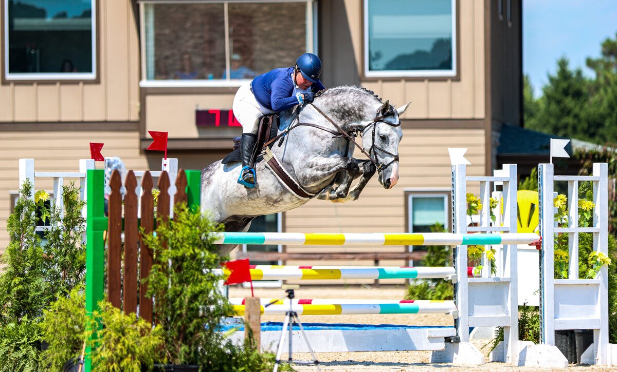 A dappled grey horse jumping a large oxer during an event at the Carolina Horse Park in Raeford, North Carolina.