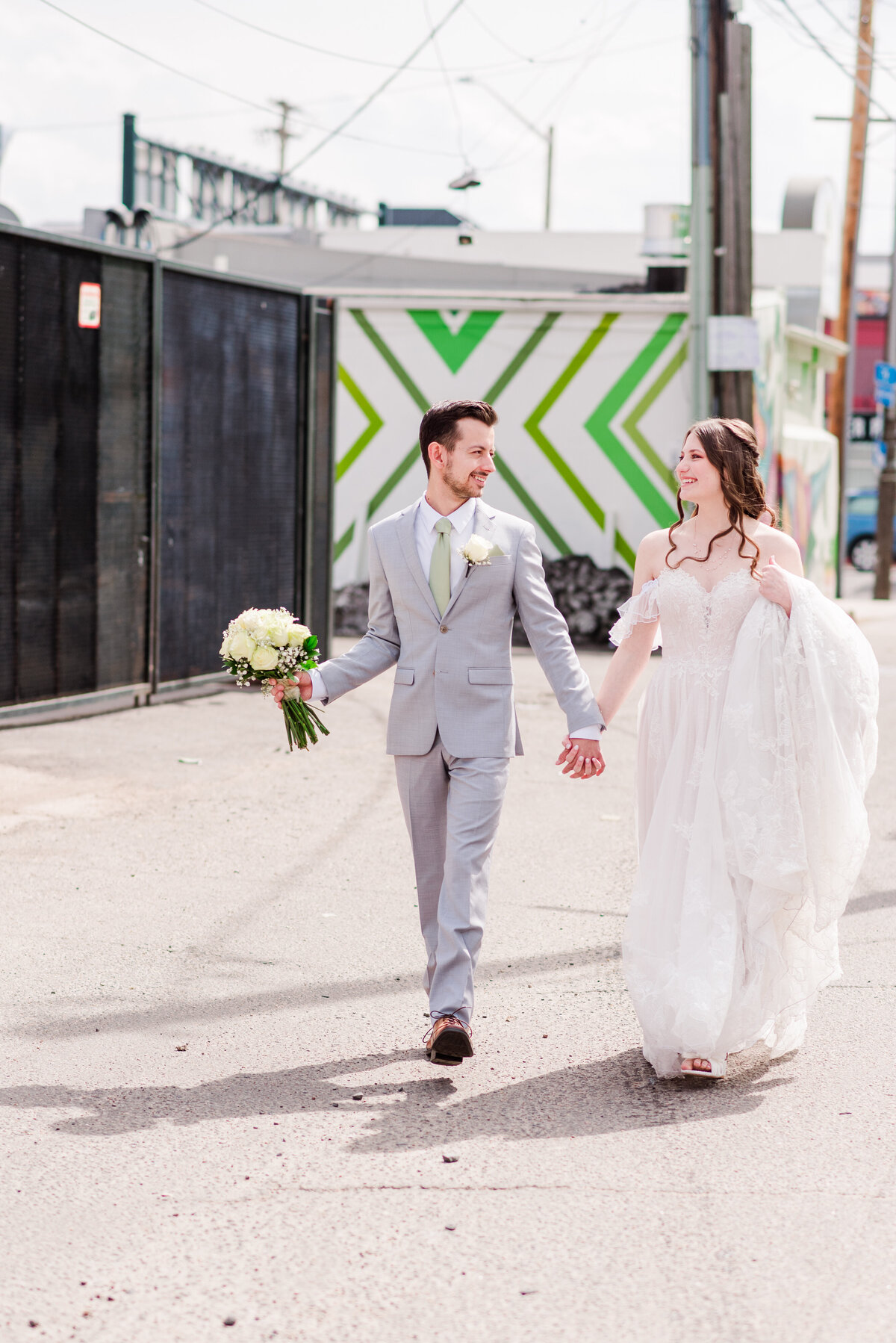 A bride and groom walk in an alleyway with vivid green grafitti walls in the background while the groom holds the flowers and the bride holds her dress and they both hold hands in the middle