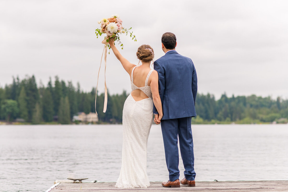 Bride-and-groom-celebrate-their-wedding-with-a-big-cheers-and-bouquet-in-the-air-as-they-look-out-at-the-lake-Venue-Green-Gates-at-Flowing-Lake-in-snohomish-photo-by-Joanna-Monger-Photography
