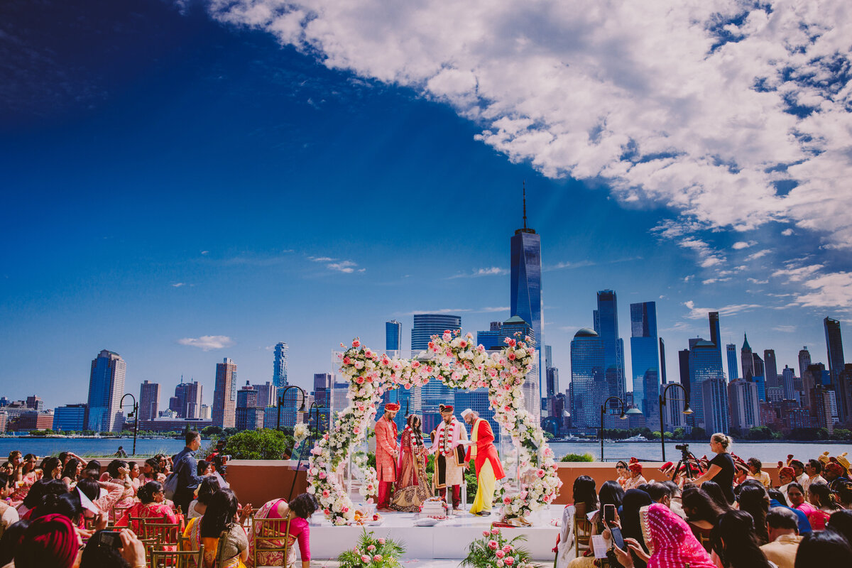 An indian bride and groom during their hindu ceremony outdoors under a glass floral mandap with the nyc skyline in the background